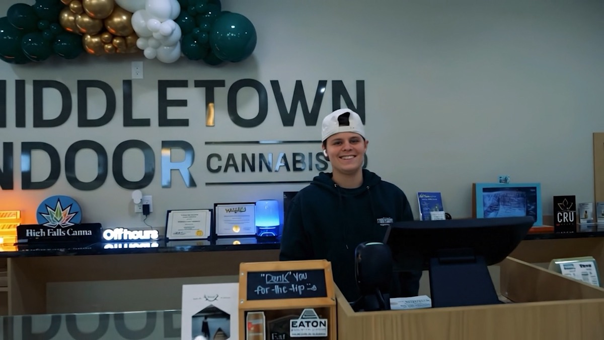 A smiling budtender at the register in front of the Middletown Indoor Cannabis Co logo wall.