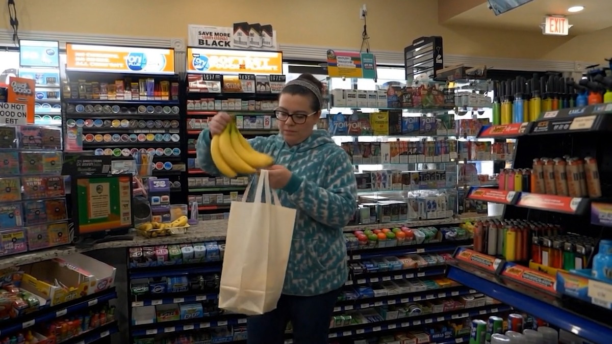 A shopper picking up bananas in a Welcome Food Mart aisle lined with snacks and drinks.